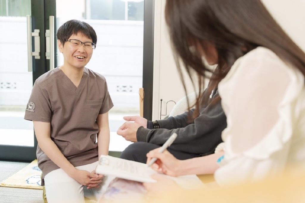 不妊鍼灸のカウンセリングイメージ。明るい室内で、茶色のスクラブを着た施術者が笑顔で話を聞き、相談者がメモを取る様子。