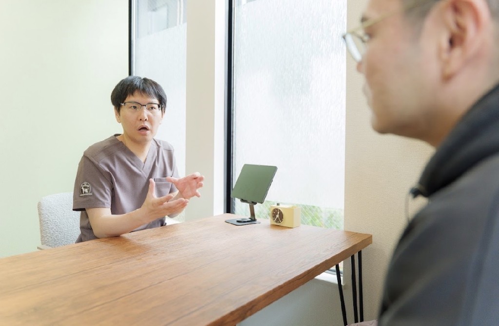 デスク越しに手を動かしながら丁寧に説明する施術者と、話を聞く相談者。明るい相談室でのカウンセリング風景。