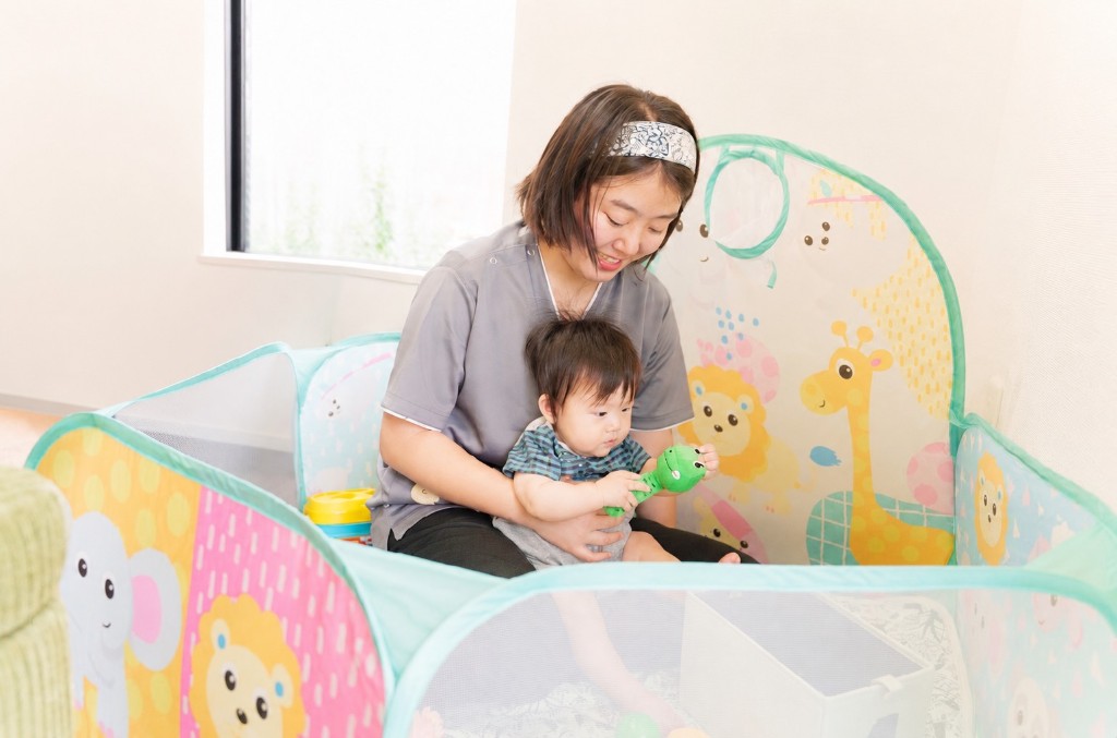 プレイヤードの中で赤ちゃんにおもちゃを渡しながら見守るスタッフ。動物柄のキッズスペースでの様子。